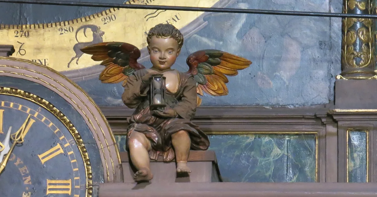 A painted wooden winged figure sits holding an hourglass next to a clock dial with gold Roman numerals